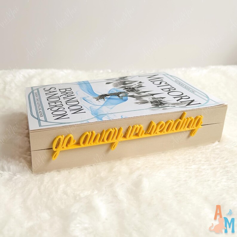 May include: A hardback book titled "Mistborn" by Brandon Sanderson, featuring a blue and white cover design. A yellow, cursive-font bookmark reads "go away I'm reading" and is placed on the book's edge. The book rests on a white, fluffy surface.