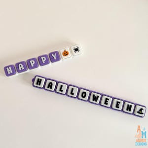 May include: Two rows of square, purple and white silicone beads. The top row spells HAPPY with a pumpkin and spider icon. The bottom row spells HALLOWEEN with a witch hat icon at the end. Each bead features a raised letter or graphic design.