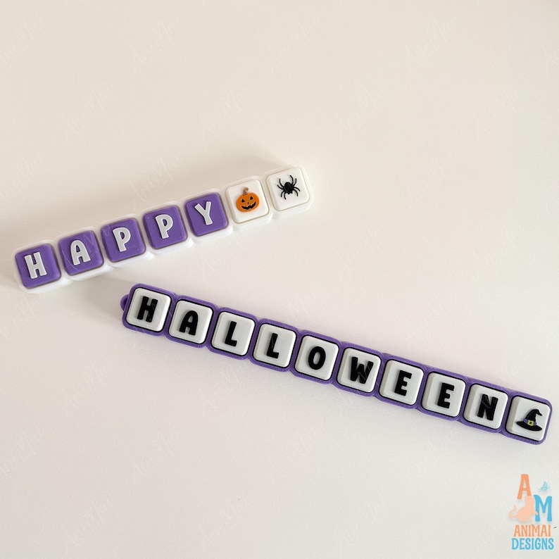 May include: Two rows of square, purple and white silicone beads. The top row spells HAPPY with a pumpkin and spider icon. The bottom row spells HALLOWEEN with a witch hat icon at the end. Each bead features a raised letter or graphic design.