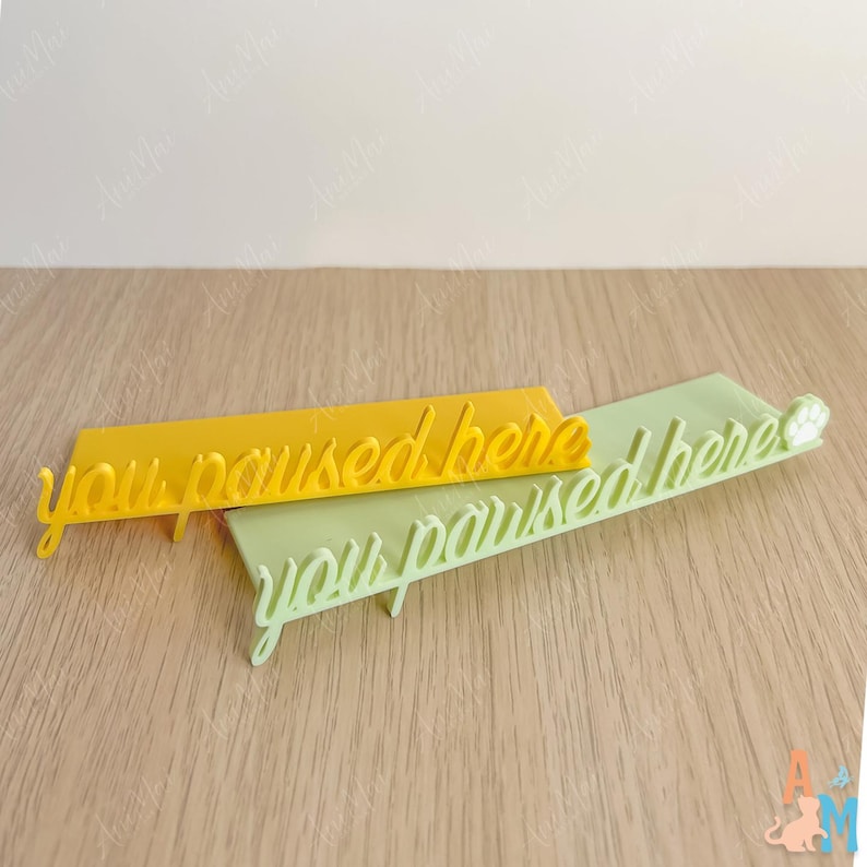 K&ouml;nnte beinhalten: Zwei rechteckige, 3D-gedruckte Schilder mit dem Text "you paused here". Eines ist gelb, das andere hellgr&uuml;n mit einem Pfotenabdruck am Ende. Die Schilder liegen auf einer hellbraunen Holzoberfl&auml;che.