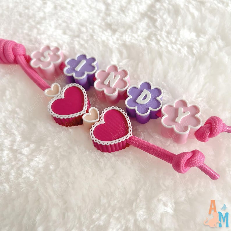 Puede incluir: Una pulsera de cord&oacute;n rosa con cuentas en forma de flor en rosa claro y morado, cada una con las letras blancas S, I, N, D, Y. El dise&ntilde;o incluye dos cuentas en forma de coraz&oacute;n rosa oscuro con bordes festoneados blancos y dos corazones peque&ntilde;os.
