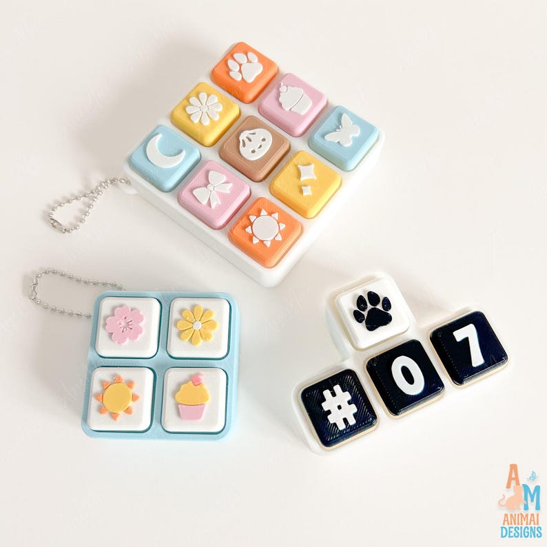 Op de afbeelding: Drie siliconen accessoires in toetsenbordstijl met pastelkleurige en donkere kleurenschema's. E&eacute;n vierkant stuk bevat negen iconen, waaronder een poot, bloem, maan, strik, zon en vlinder. Twee andere stukken tonen extra iconen en de tekens #, 0, 7.