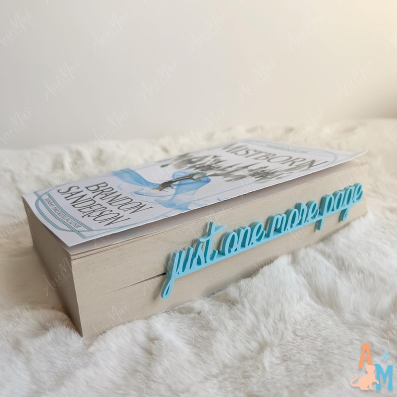 K&ouml;nnte beinhalten: Ein Buch mit dem Titel "Mistborn" von Brandon Sanderson mit einem hellblauen Lesezeichen mit der Aufschrift "just one more page". Das Buch hat ein hellblaues und wei&szlig;es Cover-Design. Das Lesezeichen besteht aus hellblauem Material.