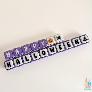 May include: A set of square, purple-bordered tiles arranged in two rows spelling HAPPY HALLOWEEN. The tiles feature white letters, a small orange jack-o'-lantern, a black spider, and a black witch hat icon on a white background.