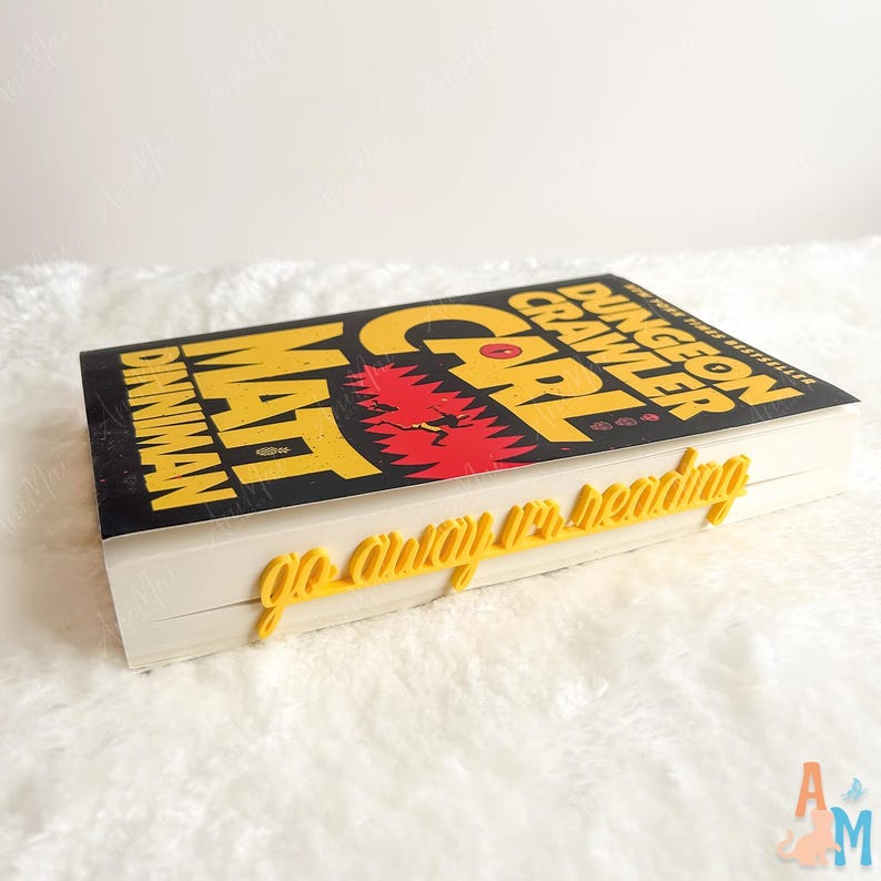 May include: A hardback book titled "Carl Dungeon Crawler" by Matt Dinniman. The cover is black with yellow lettering and a red graphic. A yellow bookmark that says "go away I'm reading" is placed on the book.