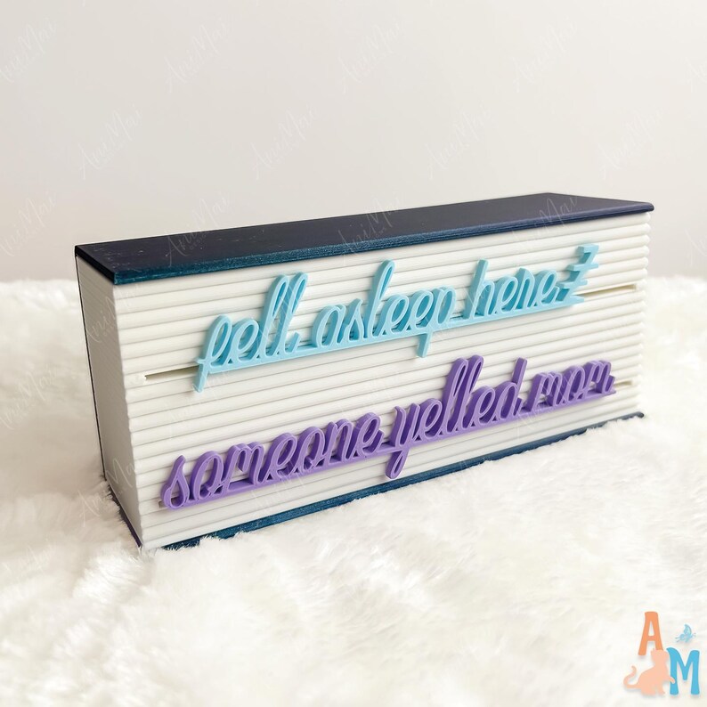 May include: A white rectangular box with a dark blue top and base. The box has horizontal lines and features the phrases "fell asleep here" in light blue and "someone yelled my" in purple, in a cursive font.