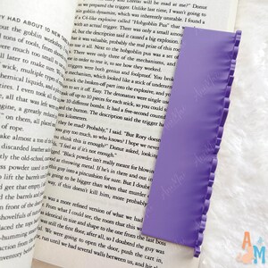 May include: A lilac bookmark with a frilled edge, lying on an open book. The bookmark is rectangular and made of a flexible material. The book's pages display printed text.