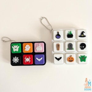 May include: Two rectangular silicone cases with attached ball chains. The black case features six colorful squares with white Halloween icons: a monster, ghost, cauldron, spiderweb, spider, and bat. The white case has nine white squares with icons.