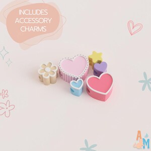 Puede incluir: Una colecci&oacute;n de cuentas de silicona coloridas en varias formas, incluyendo una flor, un coraz&oacute;n grande rosa claro, un coraz&oacute;n peque&ntilde;o azul, una estrella amarilla, un coraz&oacute;n morado y un coraz&oacute;n grande rosa oscuro. El texto INCLUDES ACCESSORY CHARMS es visible.