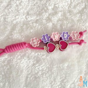 Puede incluir: Un llavero de cord&oacute;n rosa con cuentas en forma de flor en colores alternos morado y rosa, cada una con una letra blanca para deletrear el nombre SINDY. Dos dijes en forma de coraz&oacute;n rosa oscuro con bordes festoneados blancos.