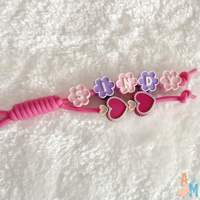Puede incluir: Un llavero de cord&oacute;n rosa con cuentas en forma de flor en colores alternos morado y rosa, cada una con una letra blanca para deletrear el nombre SINDY. Dos dijes en forma de coraz&oacute;n rosa oscuro con bordes festoneados blancos.