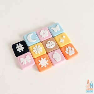 Op de afbeelding: Een verzameling van elf vierkante plastic bedels in diverse pastelkleuren, elk voorzien van een wit reli&euml;f-icoon, waaronder een maansikkel, bloem, vlinder, hashtag, knoedel, sterretjes, strik, zon, cupcake en een pootafdruk.