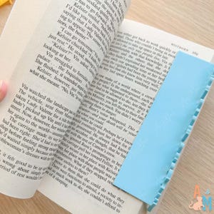 K&ouml;nnte beinhalten: Ein hellblaues Lesezeichen mit gewelltem Rand liegt auf den Seiten eines ge&ouml;ffneten Buches. Das Buch ist aufgeschlagen und zeigt gedruckten Text. Das Lesezeichen ist rechteckig und aus flexiblem Material.