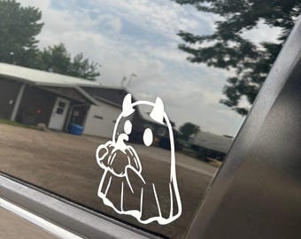 May include: White vinyl decal of a cartoon ghost with horns and a pumpkin. The decal is applied to a car window, with a rural landscape visible in the background.