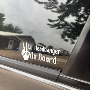 May include: White decal on a car window with the text "Lil' Headbanger On Board" in a gothic font. The decal features a skeleton hand giving the peace sign. The background shows a cloudy sky and trees.