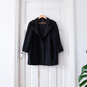 May include: A dark grey wool coat with a single button fastening and a slightly puffed shoulder detail. The coat is hanging on a wooden hanger against a white door.