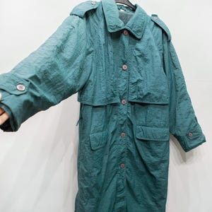 Peut inclure: Un long manteau de couleur sarcelle avec une surface texturée. Le manteau présente un col, des boutons et deux poches avant. Le manteau est suspendu à un cintre noir.