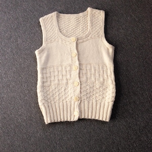 May include: A sleeveless, cream-colored knit vest featuring a scoop neckline and a front button closure with six matching buttons. The garment displays a variety of textured stitch patterns, including ribbed sections and a ribbed hemline.