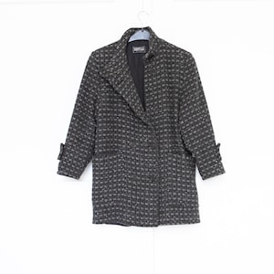 May include: A dark grey and black checked blazer featuring a notched lapel, double-breasted button closure, and three-quarter length sleeves with cuff details. The interior label reads KESTILA. The garment is constructed with a textured fabric.