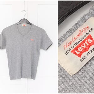 Pode incluir: Camiseta cinza de gola V com um pequeno logotipo vermelho Levi's no peito esquerdo. A etiqueta diz "The Original Levi Strauss & Co. Levi's® San Francisco."