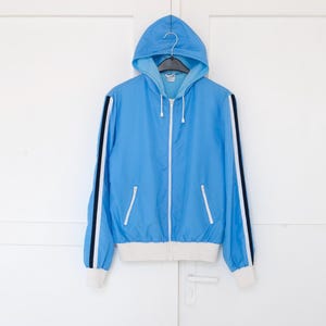 Peut inclure: Veste à capuche bleu clair avec fermeture éclair et poignets blancs. La veste a des rayures noires et blanches sur les manches. Elle est suspendue à un cintre noir sur fond blanc.
