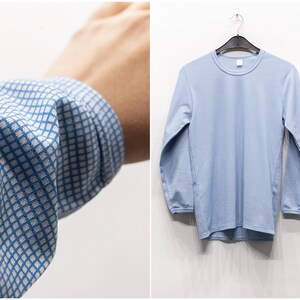Può includere: Camicia azzurra a maniche lunghe con girocollo, appesa a una gruccia nera. La camicia presenta un sottile motivo a righe. Il polsino di una camicia con un motivo a quadri blu e bianchi è visibile a sinistra.
