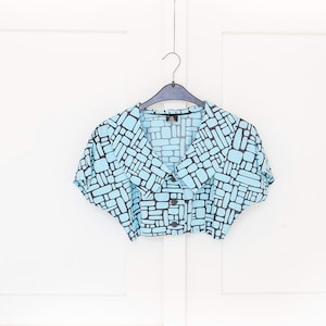 Könnte beinhalten: Hellblaues Crop-Top mit schwarzem geometrischem Muster. Das Oberteil hat kurze Ärmel, einen Kragen und wird mit drei Knöpfen geschlossen. Es hängt an einem schwarzen Kleiderbügel vor weißem Hintergrund.