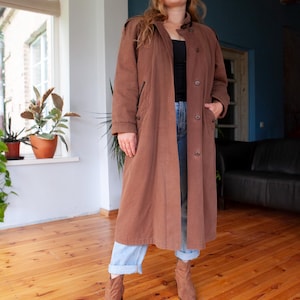 May include: A long brown coat with a button closure and pockets. The coat is worn over a black top and blue jeans.