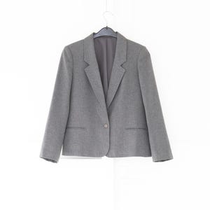 May include: A grey blazer featuring a single-button closure, notched lapels, and two horizontal welt pockets at the waist. The garment is constructed with long sleeves and a structured shoulder design, hanging from a black plastic hanger.