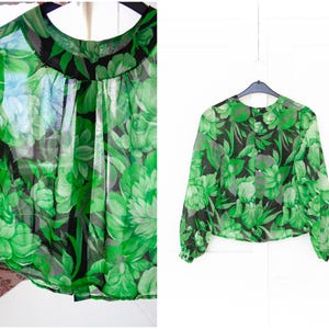May include: A sheer green blouse with a floral design in shades of green and black. The long-sleeved top is hanging on a black hanger against a white backdrop. The blouse has a round neckline.