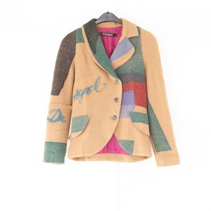 Könnte beinhalten: Ein beigefarbener Blazer mit Patchwork-Design aus strukturierten grünen, braunen, roten und blauen Stoffbahnen. Die Jacke hat ein steigendes Revers, drei Knöpfe, zwei Pattentaschen und ein leuchtend pinkes Innenfutter. Das Wort Desigual ist blau gestickt.