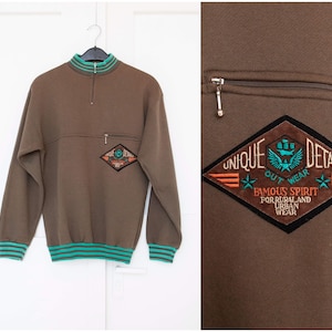 Puede incluir: Sudadera marrón con cuello alto y cremallera. La sudadera tiene una franja verde alrededor del cuello y los puños. La sudadera tiene un parche en la parte delantera que dice "Unique Details Out Wear Famous Spirit For Rural and Urban Wear".