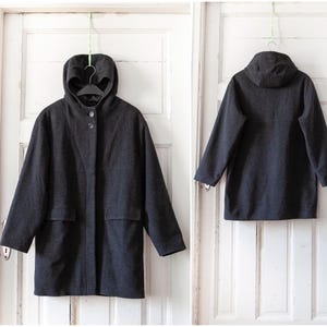 May include: A dark gray hooded coat with a button-up front and two front pockets. The coat is hanging on a black hanger from a green string, showcasing the front and back views. The coat has a simple, classic design.