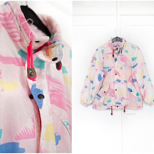 May include: A vintage pink jacket with a colourful abstract pattern. The jacket features a high collar, black buttons, and a drawstring at the waist. The design includes blue, yellow, and pink shapes. The jacket is hanging on a black hanger.