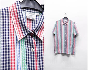 Può includere: Una camicia a maniche corte con un motivo a quadri colorati. La camicia presenta strisce verticali rosse, verdi, blu e bianche e un colletto classico. È appesa a una gruccia nera su sfondo bianco.