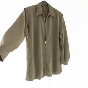 Peut inclure: Une veste à manches longues vert olive dotée d'une fermeture éclair intégrale sur le devant, d'un col pointu et de poignets boutonnés. La veste présente une texture lisse et mate, suspendue à un cintre noir sur fond blanc.