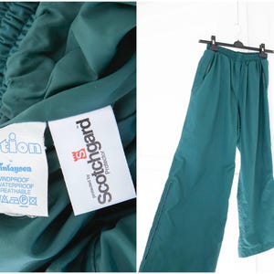May include: Teal-colored athletic pants with an elastic waistband and side pockets. The pants are hanging on a black hanger against a white background. The image also shows a close-up of the fabric with tags that read "action" and "Scotchgard Protector."