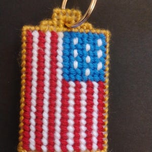 May include: A handmade keychain featuring a stitched American flag design. The flag has red and white stripes, a blue field with white stars, and a gold-colored border. The keychain includes a metal ring for attaching keys.