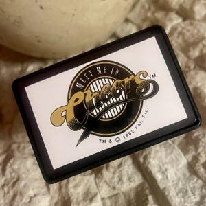 May include: A rectangular black magnet with the words "Meet Me In Cheers" in gold script. The magnet has a white background with a black and gold logo. The copyright date is 1992.