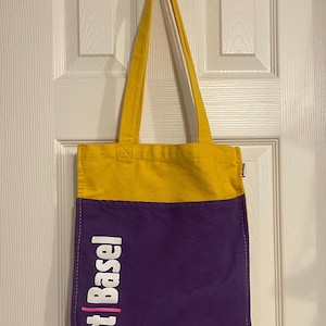 May include: A two-tone tote bag with a yellow top and purple bottom. The words "Art Basel" are printed vertically in white with a pink vertical line on the left side. The bag has yellow straps and is hanging on a metal hook.