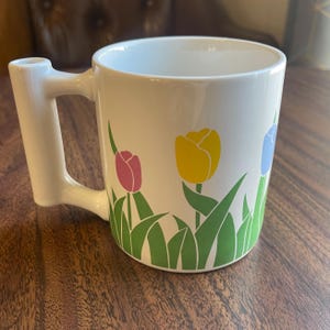 May include: White ceramic mug with a unique handle shaped like a pipe. The mug features a colorful tulip design with yellow, pink, and blue tulips on a green stem.