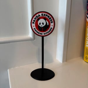 Könnte beinhalten: Ein schwarzer Ständer mit einem rot-weißen Panda Express-Logo, das einen schwarz-weißen Pandabären zeigt. Das Logo lautet "Panda Express Chinese Kitchen".