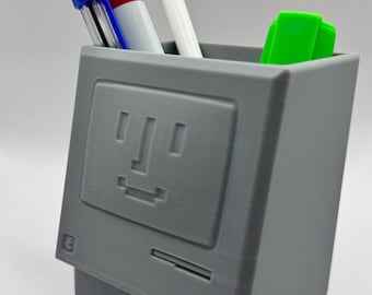 May include: A gray desktop computer-shaped pencil holder with a simple smiley face on the front. The holder has a blue pen, a red marker, a white marker, and a green highlighter inside.