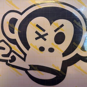 Op de afbeelding: Een zwart-witte aapgezichtsticker. Het apengezicht heeft een boze uitdrukking met een X over één oog en een schuine streep over het andere. De sticker meet 28 cm bij 21,6 cm.