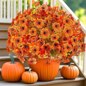May include: An arrangement of orange flowers in a matching pumpkin-shaped pot, with three pumpkins of varying sizes. The pumpkins and pot are a vibrant orange, creating a festive autumn display. The flowers have dark centers.