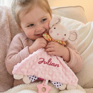 Puede incluir: Una manta de seguridad rosa para bebé con una cabeza de conejo de peluche y el nombre "Julian" bordado en la parte delantera. La manta tiene una superficie texturizada y detalles de lunares. Un mordedor rosa en forma de estrella está adjunto.