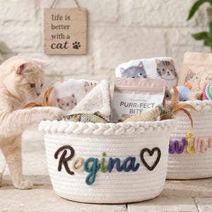 Puede incluir: Dos cestas de cuerda blanca llenas de juguetes y golosinas para gatos. Una cesta tiene el nombre "Regina" y un corazón. La otra cesta tiene el nombre "Winnie". Un gato interactúa con una de las cestas. Un letrero dice "la vida es mejor con un gato".