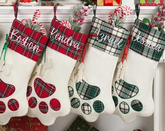 May include: Four white Christmas stockings shaped like paw prints, each with a plaid cuff and personalized name. The stockings have red and green plaid accents and are hanging on a white mantelpiece, decorated for the holidays.