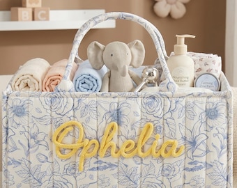 Peut inclure: Un panier cadeau pour bébé avec un motif floral bleu et blanc, contenant des couvertures roulées, un éléphant en peluche, de la lotion et un hochet. Le nom "Ophelia" est écrit en jaune.