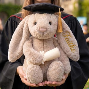 Puede incluir: Un conejo de peluche con birrete y diploma enrollado. El conejo es de color marrón claro con orejas largas y el nombre "Faithe" bordado en una oreja. El birrete es negro con una borla dorada.